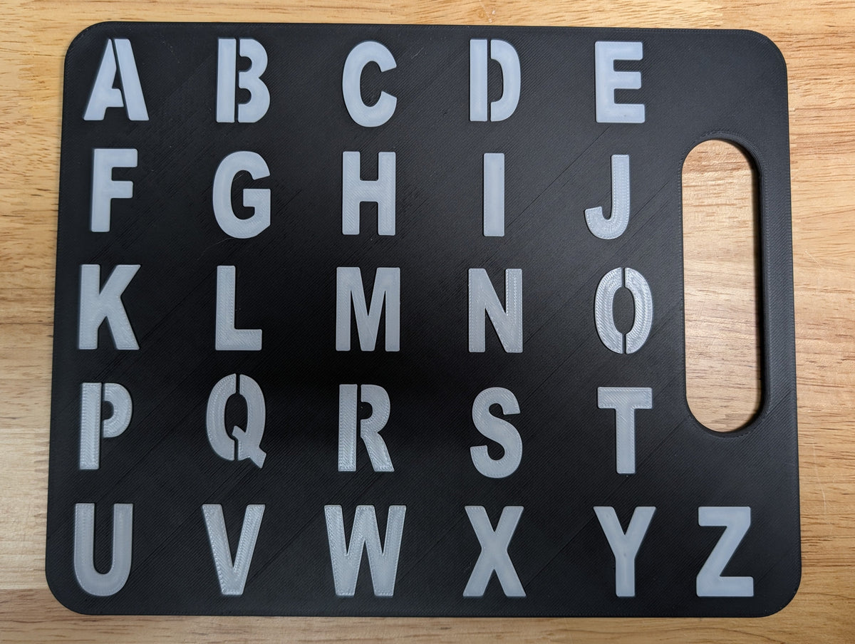 A black rectangular alphabet board with raised silver letters A through Z and a handle cutout on the right side, placed on a light wood surface.
