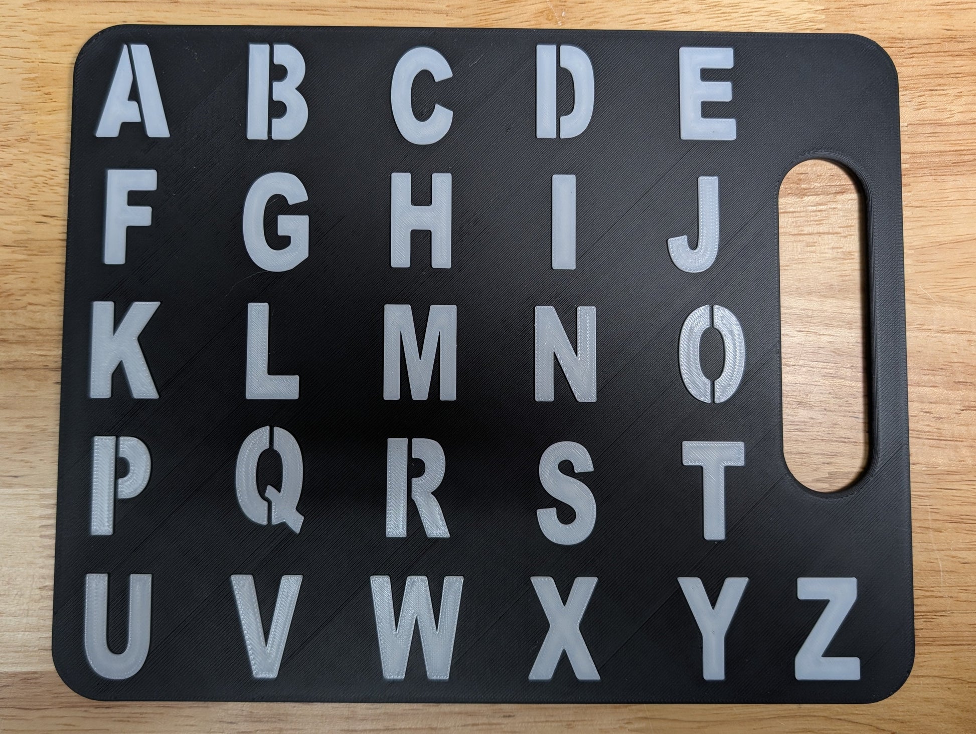 A black rectangular alphabet board with raised silver letters A through Z and a handle cutout on the right side, placed on a light wood surface.