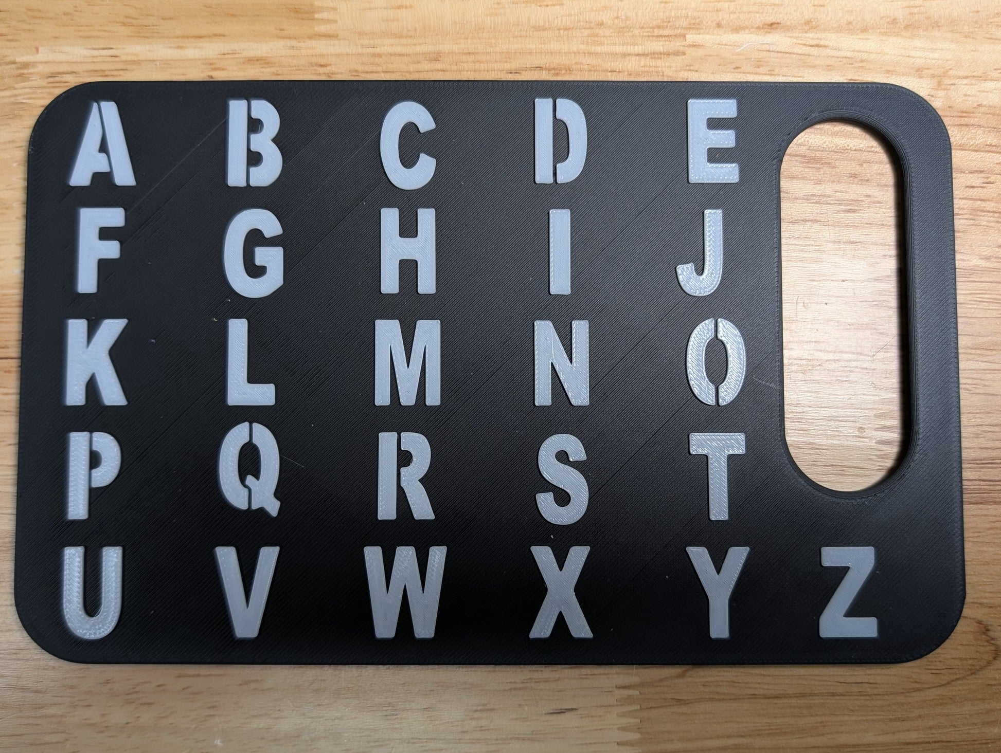 A black alphabet board with raised silver letters A through Z and a rounded rectangular cutout at the bottom, placed on a light wood surface.