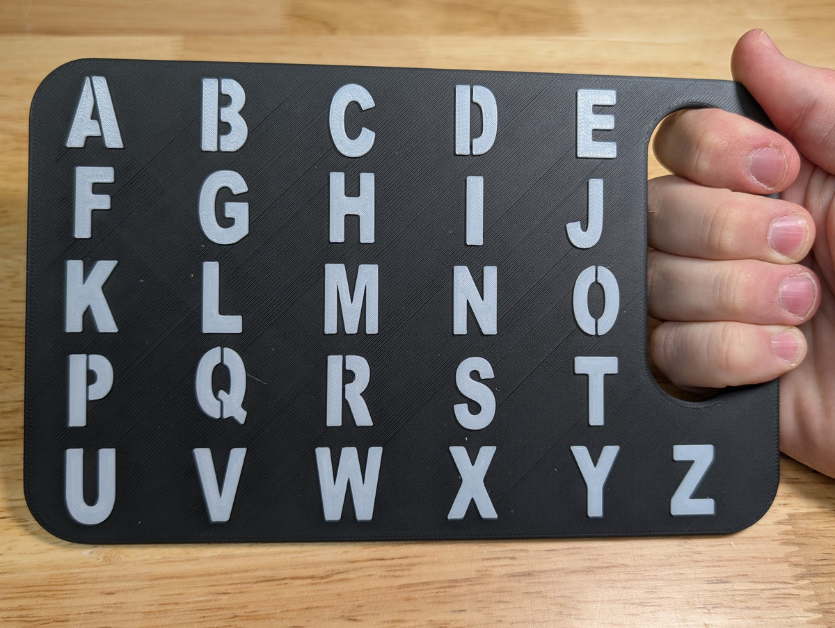 A hand holding a black alphabet learning board with white uppercase letters A through Z on a light wood tabletop.