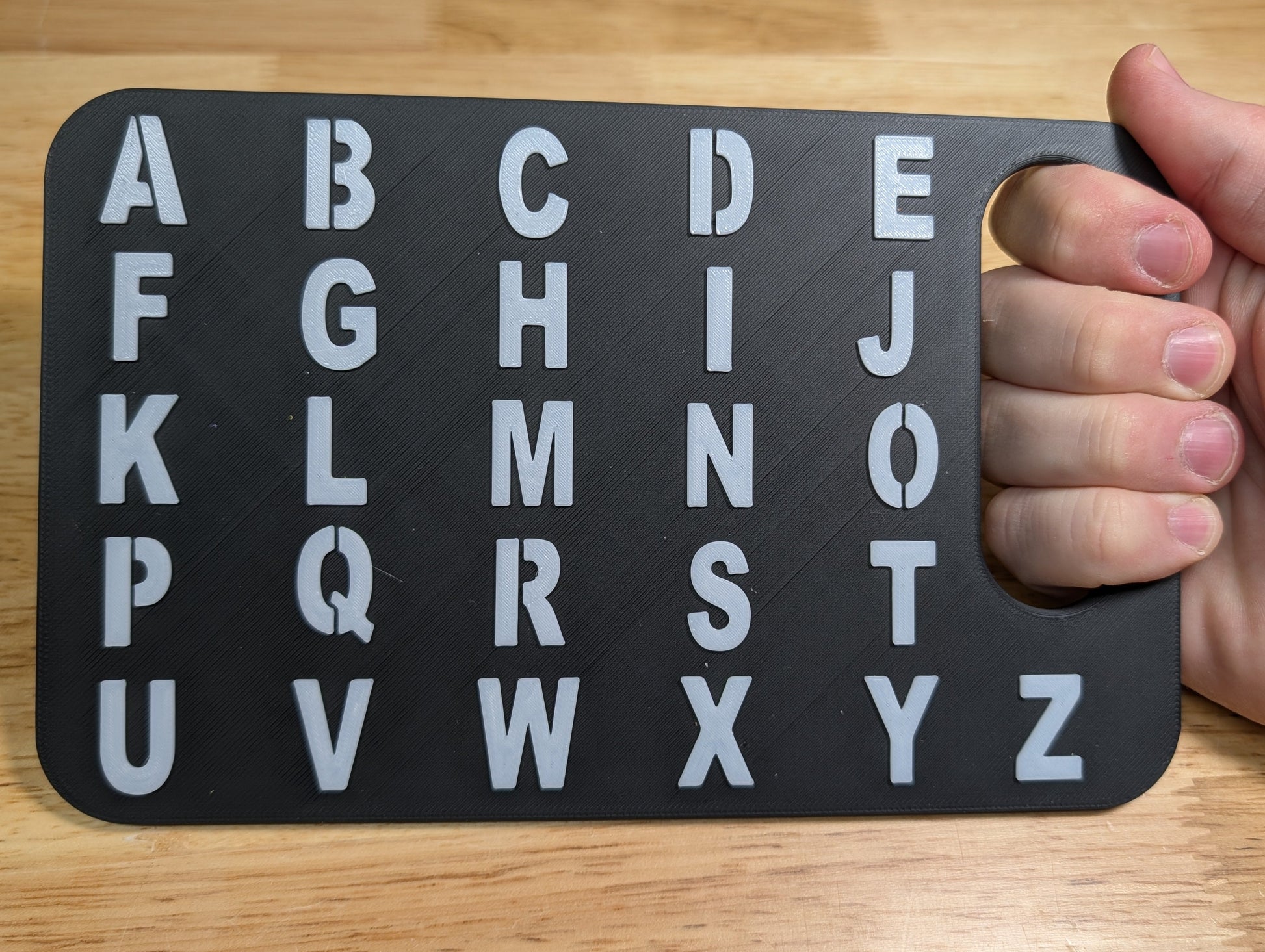 A hand holding a black alphabet learning board with white uppercase letters A through Z on a light wood tabletop.