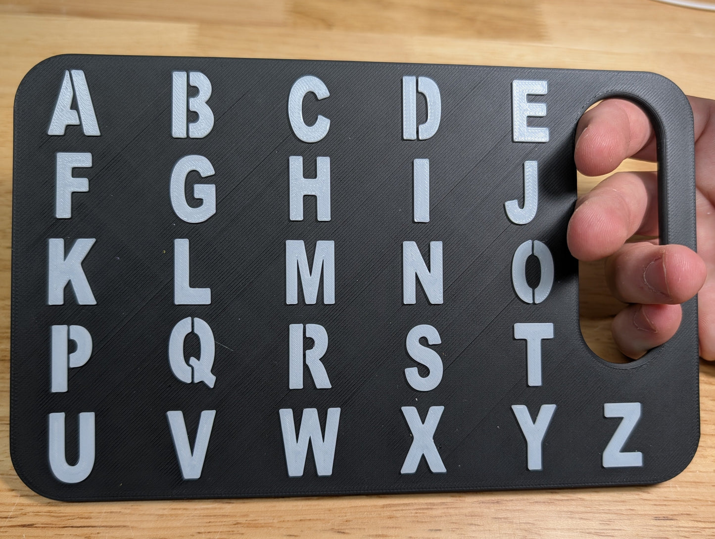 A black rectangular alphabet learning board with raised white letters A through Z and a handle cutout on the right side, held by a hand over a light wood tabletop.